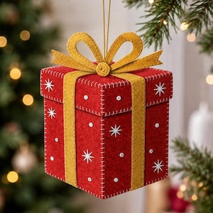 May include: A red felt gift box ornament featuring a gold felt ribbon and bow on top. The box is decorated with white embroidered star patterns and small white dots, with visible white stitching along the edges and a gold hanging string.