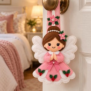 May include: A felt angel ornament hanging from a door handle. The angel has brown hair with a pearl headband, a pink dress with a bow, and white wings. It is decorated with small green holly leaves, red berries, and pink poinsettia flowers.