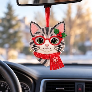 Pode incluir: Um ornamento de feltro em forma de gato pendurado num espelho retrovisor. O gato cinzento tigrado tem olhos verdes grandes, óculos vermelhos, um cachecol vermelho com um padrão de floco de neve branco e um azevinho verde com bagas vermelhas.