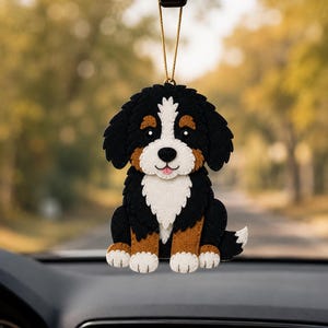 Peut inclure: Ornement de voiture en feutre représentant un Bouvier Bernois. Le chien est noir, blanc et marron, avec un visage souriant et une posture assise. Il est suspendu à une ficelle dorée, devant un arrière-plan extérieur flou.