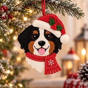 Puede incluir: Un adorno de fieltro con forma de perro de montaña de Berna que lleva un gorro de Papá Noel rojo con borde blanco, hojas de acebo verdes y bayas rojas. El perro luce una bufanda roja decorada con un patrón de copo de nieve blanco bordado.