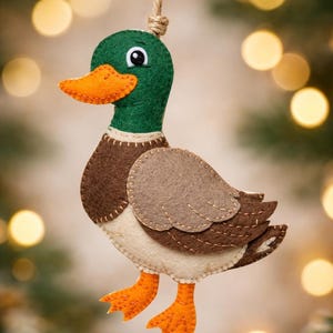 May include: A handmade felt duck ornament with a green head, orange beak and feet, and brown and tan body. The duck is suspended by a jute twine loop, perfect for holiday decor. The ornament is approximately 5 inches tall.