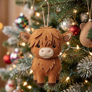 Puede incluir: Adorno navideño de fieltro marrón con forma de vaca de las Tierras Altas, con detalles cosidos, hocico claro y ojos negros. El adorno cuelga de una cuerda de hilo natural, expuesto en un árbol de Navidad decorado.