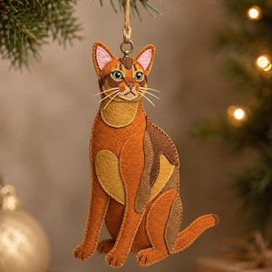 Könnte beinhalten: Ein Katzen-Anhänger aus Filz in Orange-, Braun- und Beigetönen mit gestickten Details, grünen Augen und rosafarbenen Ohrinnenseiten. Die Katze hängt an einer Juteschnur, die an einem Metallring am Kopf befestigt ist und zeigt geschichteten Stoff.