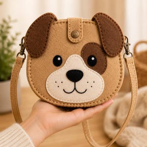 Puede incluir: Un bolso bandolera de fieltro color canela con forma de cara de perro, orejas marrón oscuro, hocico color crema, ojos negros, nariz negra y un pequeño cierre de presión metálico. El bolso incluye una correa de hombro de fieltro a juego.