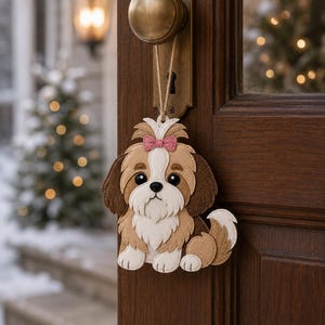 May include: A felt Shih Tzu dog ornament with a pink bow, hanging from a door knob. The ornament features brown, white, and tan colors. The background shows a snowy outdoor scene with a Christmas tree.