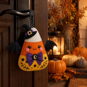 May include: A felt candy corn ornament hanging from a door handle, featuring a black witch hat, purple bow with a crescent moon charm, and black bat wings. The candy corn has a white top, orange middle, and yellow bottom with embroidered stars.