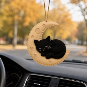Puede incluir: Un adorno colgante para coche con un gato de fieltro negro durmiendo sobre una luna creciente de fieltro beige. La luna tiene hendiduras circulares y está suspendida por un cordel marrón fino. El gato tiene ojos, nariz y bigotes bordados.