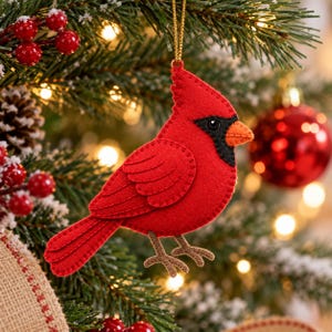 Op de afbeelding: Een vogelornament van vilt in de vorm van een kardinaal, uitgevoerd in felrood met een zwart gezichtsmasker en een oranje snavel. De vogel heeft zichtbare stiksels langs de vleugels en het lichaam en hangt aan een dun gouden koordje.