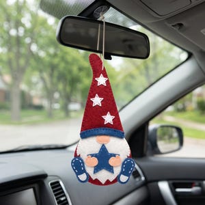 May include: A felt gnome car ornament with a red hat decorated with white stars, a blue brim, and a white beard. The gnome holds a blue star and has blue feet. The ornament hangs from a string attached to a car's rearview mirror.