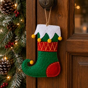 May include: A felt Christmas stocking ornament in green and red with a white scalloped cuff. The stocking features yellow pom-poms along the cuff and toe, a red band with yellow zigzag stitching, and a red heel patch, hanging from a brown twine loop.