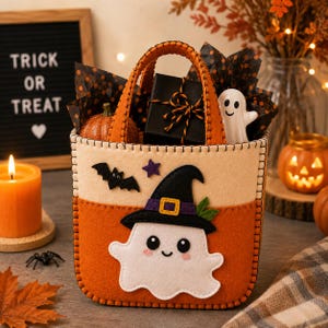 May include: A felt Halloween treat bag featuring a white ghost wearing a black witch hat with a purple band. The bag is orange and cream with stitched edges, decorated with a small black bat and a purple star. It contains a black gift box.