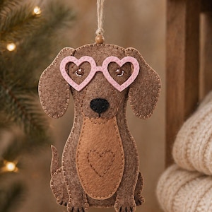 May include: A brown felt dachshund ornament wearing pink heart-shaped glasses. The dog has a lighter brown belly with a heart-shaped patch. A string is attached for hanging. The ornament is a festive decoration.