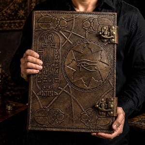 May include: A large, brown leather-bound book with detailed embossed designs, including an eye symbol and hieroglyphs. The book features a clasp closure and an aged appearance, suggesting a historical or fantasy theme.