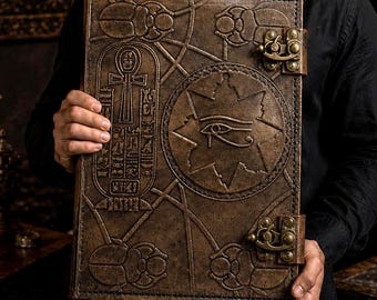 Op de afbeelding: Een rechthoekig, bruin, in leer gebonden boek met in reliëf aangebrachte Egyptische motieven, waaronder een Oog van Horus in een ster, een ankh en scarabeeën. De kaft wordt vastgezet met twee sierlijke, messingkleurige metalen sluitingen.