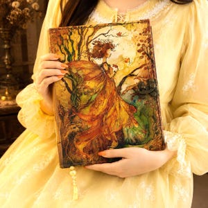 May include: A leather-bound journal with a fantasy art cover. The artwork depicts a woman in a flowing gown, trees, and a full moon. The journal has a metal clasp and a decorative tassel.