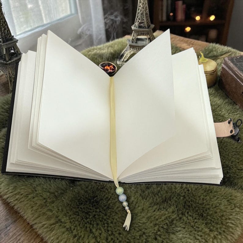 May include: An open journal with blank white pages, a yellow ribbon bookmark, and a beaded tassel. The journal has a black cover and a leather strap closure. The background includes a miniature Eiffel Tower and a green, textured surface.