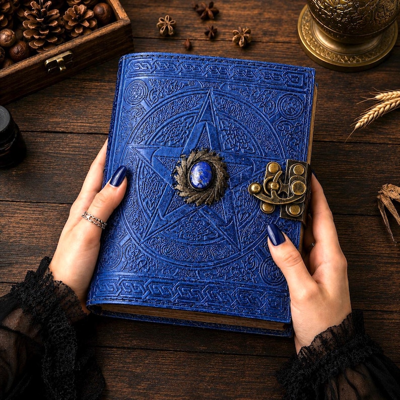 Peut inclure: Un journal en cuir bleu avec un pentagramme en relief sur la couverture. Le journal est orn&eacute; d'un fermoir d&eacute;coratif et d'une pierre centrale. Le journal est tenu par des mains avec du vernis &agrave; ongles bleu fonc&eacute;.