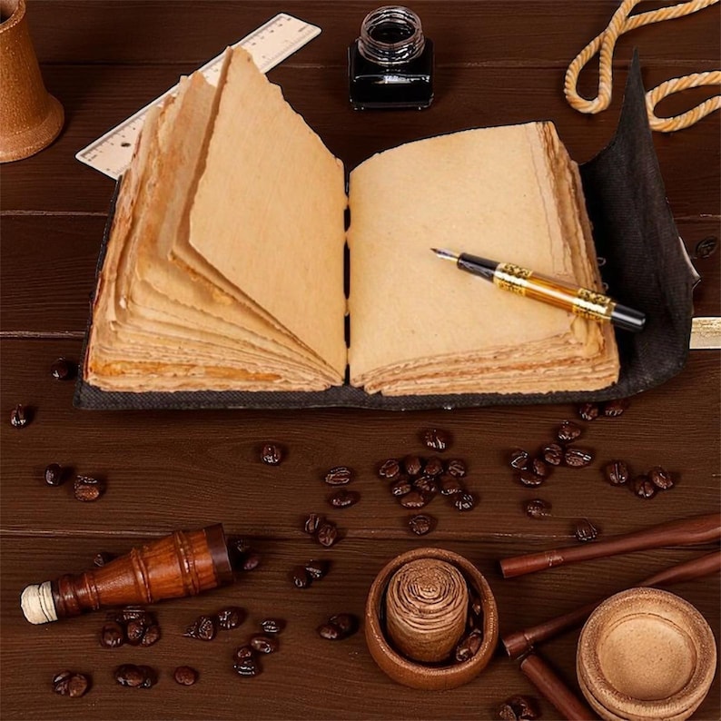 Peut inclure: Un journal ouvert aux pages &eacute;paisses et textur&eacute;es, d'aspect vieilli, pos&eacute; sur une surface en bois sombre. Un stylo or et noir repose sur la page de droite. Des grains de caf&eacute;, un petit r&eacute;cipient en bois et un encrier entourent le journal.