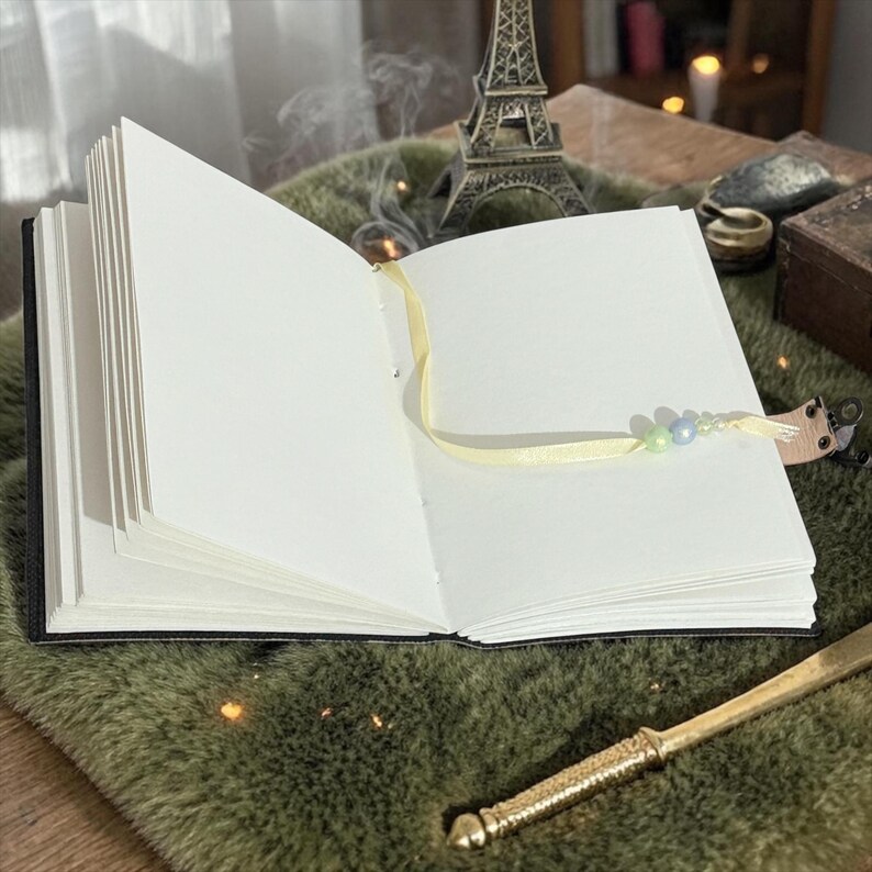 May include: An open, blank journal with a black cover and a yellow ribbon bookmark adorned with beads. The journal is on a green surface, with a miniature Eiffel Tower and a decorative letter opener nearby. The pages are crisp white.