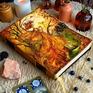May include: A decorative journal with a detailed illustration of a woman in a flowing gown, set against a backdrop of trees and a full moon. The cover features a metal clasp and is surrounded by other decorative items.