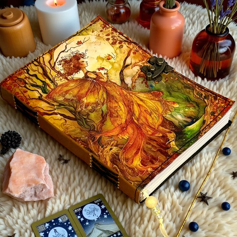 May include: A decorative journal with a detailed illustration of a woman in a flowing gown, set against a backdrop of trees and a full moon. The cover features a metal clasp and is surrounded by other decorative items.