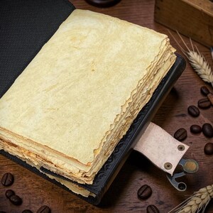 Peut inclure: Un journal ouvert de style ancien avec des pages jaunies. Le journal est reli&eacute; en cuir noir et poss&egrave;de un signet en cuir. Des grains de caf&eacute;, des crayons et des &eacute;pis de bl&eacute; sont dispers&eacute;s autour du journal sur une surface en bois.