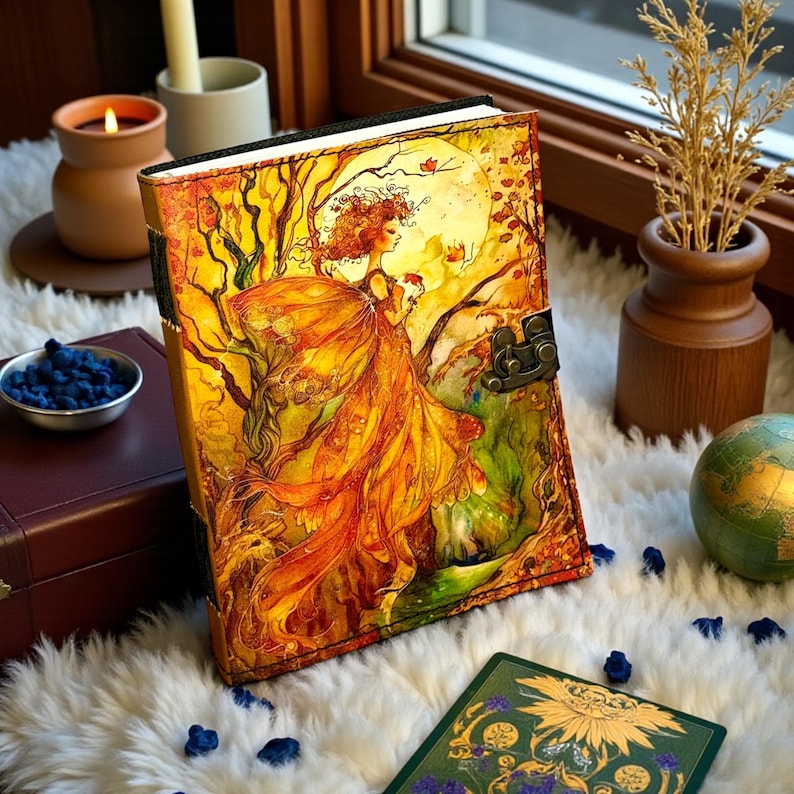 May include: A leather-bound journal with a vibrant illustration of a woman in a flowing orange dress, set against a backdrop of trees and a full moon. The journal features a metal clasp and is displayed on a fluffy white surface.