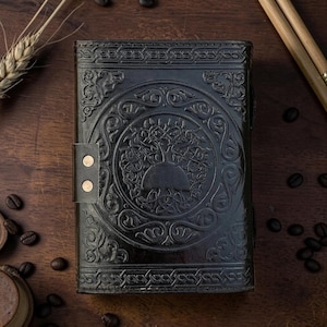 Peut inclure: Journal en cuir noir avec un motif en relief d'arbre de vie. Le journal a une fermeture &agrave; boucle avec deux rivets dor&eacute;s. La couverture pr&eacute;sente des entrelacs celtiques complexes autour de l'arbre. Le journal est ferm&eacute; et pos&eacute; sur une surface en bois.