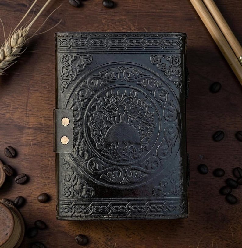Peut inclure: Journal en cuir noir avec un motif en relief d'arbre de vie. Le journal a une fermeture &agrave; boucle avec deux rivets dor&eacute;s. La couverture pr&eacute;sente des entrelacs celtiques complexes autour de l'arbre. Le journal est ferm&eacute; et pos&eacute; sur une surface en bois.