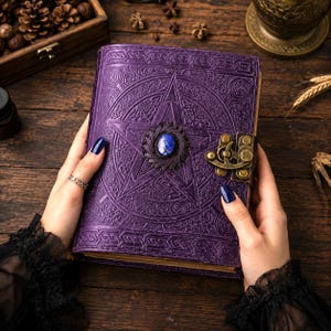 Peut inclure: Un journal en cuir violet avec un motif de pentagramme en relief et un fermoir d&eacute;coratif. Le journal pr&eacute;sente une pierre pr&eacute;cieuse bleue au centre de l'&eacute;toile. Il est tenu par des mains avec du vernis &agrave; ongles bleu fonc&eacute;.