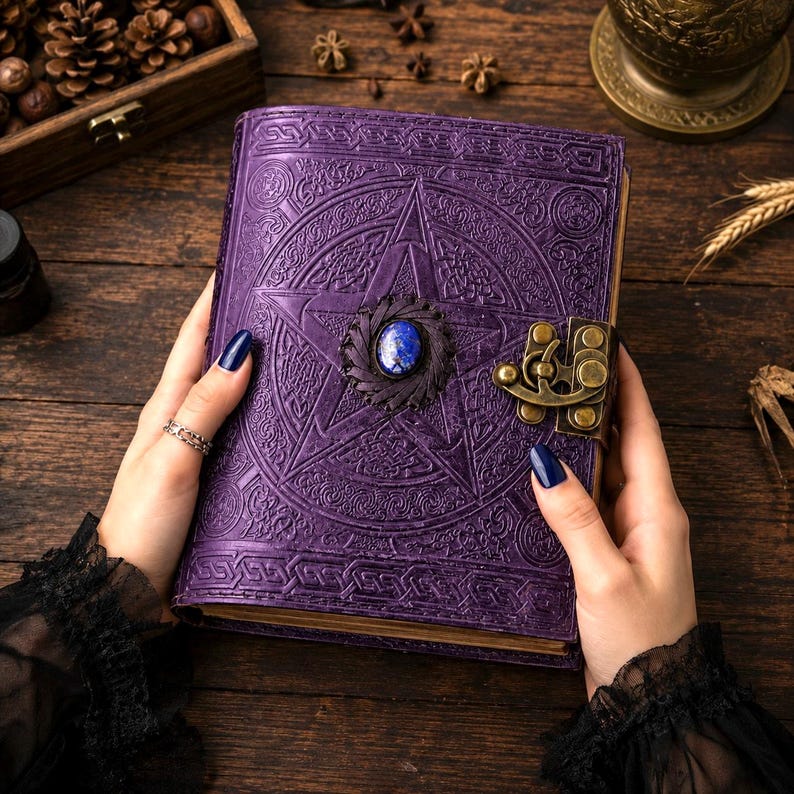 Peut inclure: Un journal en cuir violet avec un motif de pentagramme en relief et un fermoir d&eacute;coratif. Le journal pr&eacute;sente une pierre pr&eacute;cieuse bleue au centre de l'&eacute;toile. Il est tenu par des mains avec du vernis &agrave; ongles bleu fonc&eacute;.