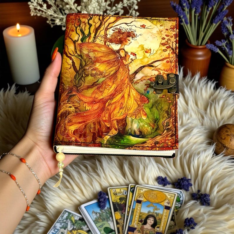 May include: A fantasy-themed journal with a detailed illustration of a fairy in autumnal colors. The cover features a clasp closure and a decorative tassel. The journal is held in a hand, with tarot cards visible in the foreground.
