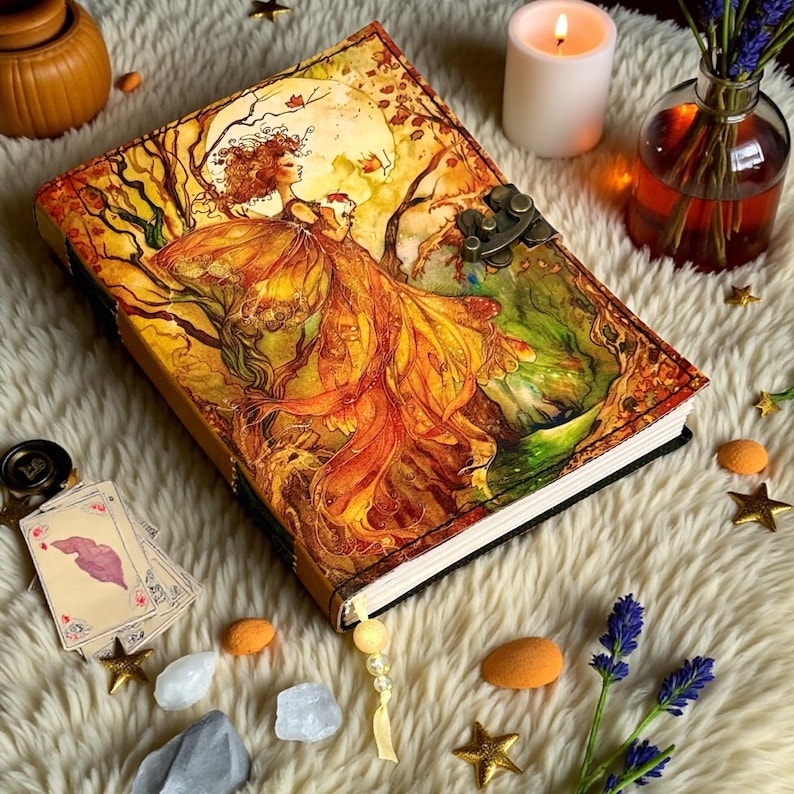 May include: A decorative journal with a detailed illustration of a woman in a flowing gown, set against a backdrop of trees and a full moon. The cover features a metal clasp. The journal is surrounded by candles, crystals, and star-shaped decorations.