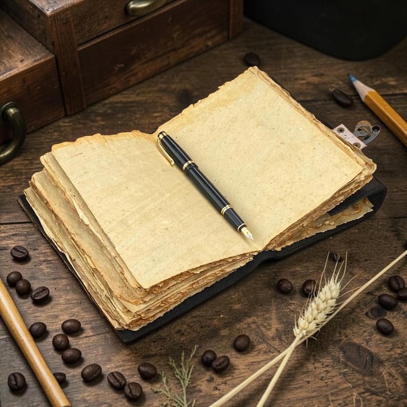Peut inclure: Un carnet ouvert avec des pages vierges et un stylo plume noir et dor&eacute;. Le carnet est pos&eacute; sur une surface en bois, entour&eacute; de grains de caf&eacute;, d'un crayon et d'&eacute;pis de bl&eacute;.
