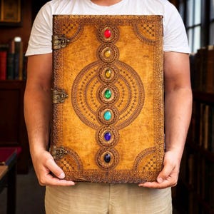May include: A large, antique-style book with a textured, tan leather cover. The cover features intricate embossed designs and is adorned with seven colorful, gemstone-like embellishments. The book has metal clasps and a detailed border.
