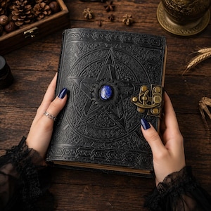 Peut inclure: Un journal en cuir noir avec un pentagramme en relief et une pierre pr&eacute;cieuse bleue au centre. Le journal est ferm&eacute; par un fermoir en laiton antique, r&eacute;v&eacute;lant un savoir-faire d&eacute;taill&eacute; et un aspect vieilli.