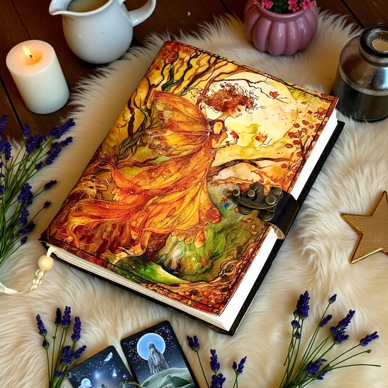 May include: A hardcover journal with a detailed illustration of a woman in a flowing orange dress, set against a backdrop of trees and a full moon. The journal has a black spine and a metal clasp. A white tassel hangs from the top.