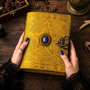 Peut inclure: Un journal en cuir jaune dor&eacute; avec un pentagramme en relief et une pierre pr&eacute;cieuse bleue au centre. Le journal a un fermoir orn&eacute; et est tenu par des mains avec du vernis &agrave; ongles bleu fonc&eacute;. La couverture est tr&egrave;s d&eacute;taill&eacute;e.