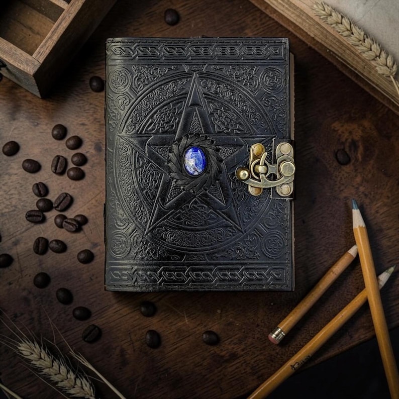 Peut inclure: Journal en cuir noir avec un pentagramme en relief et une pierre pr&eacute;cieuse bleue au centre. Le journal est dot&eacute; d'un fermoir en laiton antique. Des crayons et des grains de caf&eacute; entourent le journal, cr&eacute;ant une esth&eacute;tique vintage.