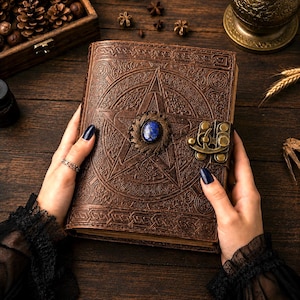 Peut inclure: Un journal en cuir marron avec un pentagramme en relief et une pierre pr&eacute;cieuse bleue. Le journal a un fermoir en m&eacute;tal et est tenu par des mains avec du vernis &agrave; ongles fonc&eacute;. Le journal est sur une surface en bois.