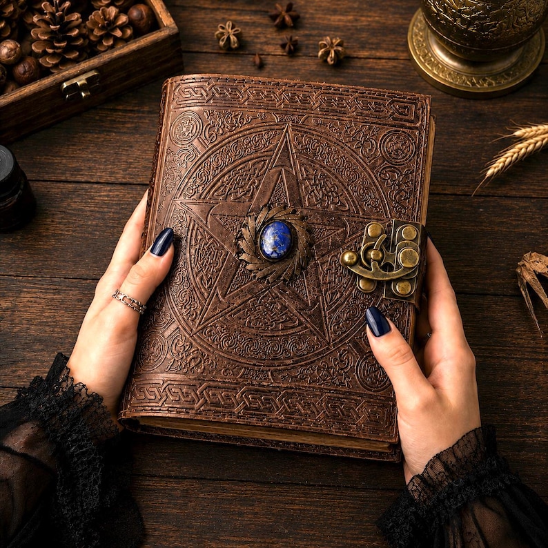 Peut inclure: Un journal en cuir marron avec un pentagramme en relief et une pierre pr&eacute;cieuse bleue. Le journal a un fermoir en m&eacute;tal et est tenu par des mains avec du vernis &agrave; ongles fonc&eacute;. Le journal est sur une surface en bois.