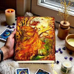 May include: A leather-bound journal with an illustration of a woman in a flowing orange dress, set against a full moon and autumnal trees. The journal is held open, revealing blank pages. Tarot cards and candles are in the background.