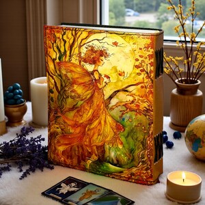 May include: A large, illustrated journal featuring a fairy in a golden dress against a full moon backdrop. The cover art is rich with autumnal colors, with a leather spine and decorative clasps. Tarot cards and candles surround the journal.