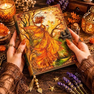 May include: A leather-bound journal with an illustration of a woman in a flowing orange dress, set against a backdrop of trees and a full moon. The journal is held open, revealing the detailed artwork. The scene is rich in autumnal colors.
