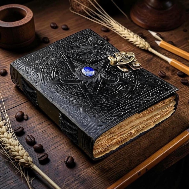 Peut inclure: Un journal en cuir noir avec un motif d'&eacute;toile en relief et une pierre pr&eacute;cieuse bleue au centre. Le journal a un fermoir en m&eacute;tal et des pages en papier vieilli. Des &eacute;pis de bl&eacute;, des grains de caf&eacute; et des crayons sont dispos&eacute;s sur une surface en bois.