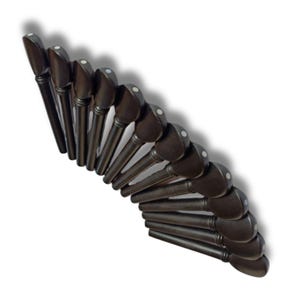 May include: A collection of dark brown violin pegs arranged in a curved pattern. Each peg has a rounded head with a small white dot and a tapered shaft. The pegs are set against a white background, with a shadow.