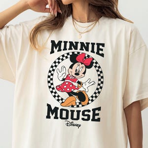 May include: Ivory-colored oversized t-shirt featuring a Minnie Mouse graphic. The graphic includes Minnie in a red polka-dot dress, a red bow, and the words "MINNIE MOUSE" in black, with "Disney" below. The shirt has short sleeves.