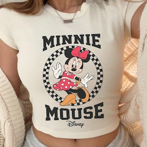 K&ouml;nnte beinhalten: Cremefarbenes Crop-Top mit Minnie Mouse-Grafik. Das Design enth&auml;lt die schwarzen W&ouml;rter "MINNIE MOUSE", mit Minnie in einem roten gepunkteten Kleid. Das Top hat einen runden Ausschnitt und kurze &Auml;rmel.