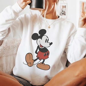 May include: White sweatshirt featuring a classic Mickey Mouse graphic. The cartoon character is depicted in black, red, and tan colors. The sweatshirt has long sleeves and a crew neck. A black mug is held up to the face.